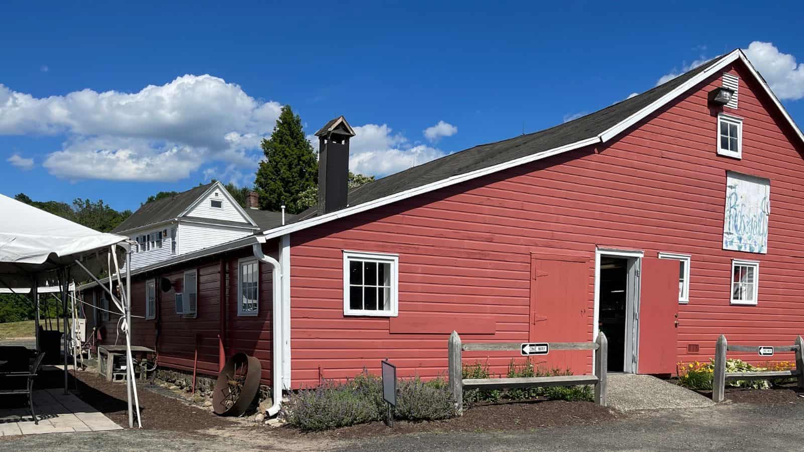A red wooden barn with a partially open door, white trim, and a small sign, next to a white house and a canopy tent under a blue sky with clouds.