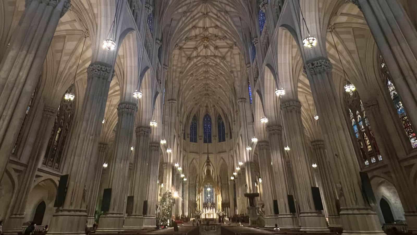 Interior view of a grand cathedral with tall columns, vaulted ceilings, hanging lights, and stained glass windows, leading to an ornate altar at the far end.