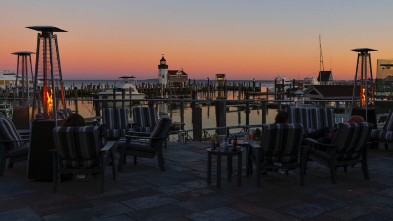 Outdoor seating area with striped chairs and patio heaters overlooks a marina with boats and a lighthouse at sunset, sky showing orange and purple hues.
