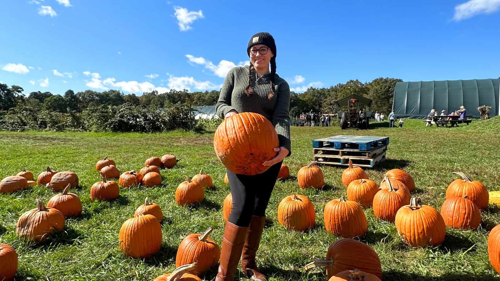 A woman wearing glasses and a beanie stands outdoors on grass, holding a large pumpkin among many pumpkins, with people and a tractor visible in the background.