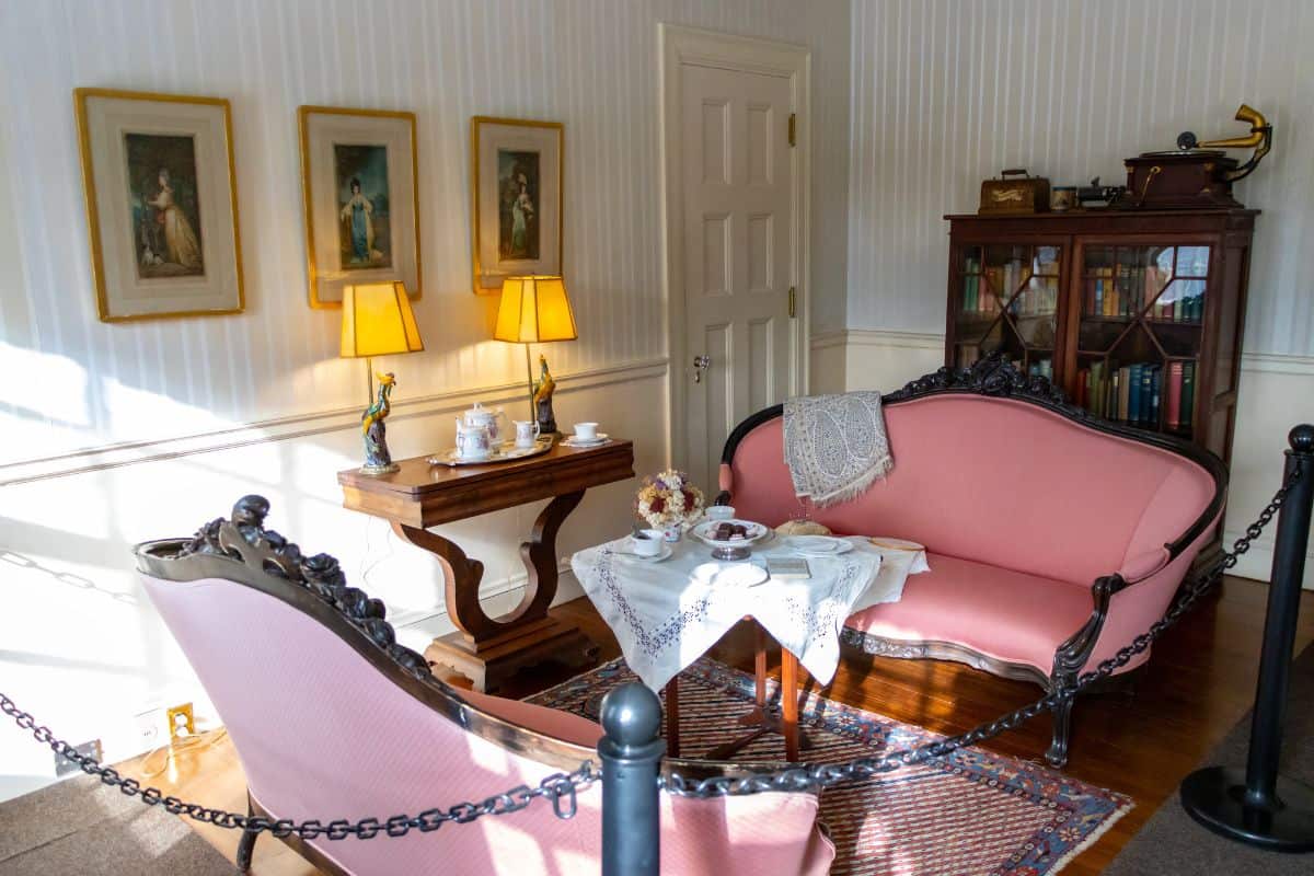 A small, vintage parlor room with pink upholstered sofas, a wooden table set for tea, a cabinet with books, and framed art on the wall, separated by a rope barrier.