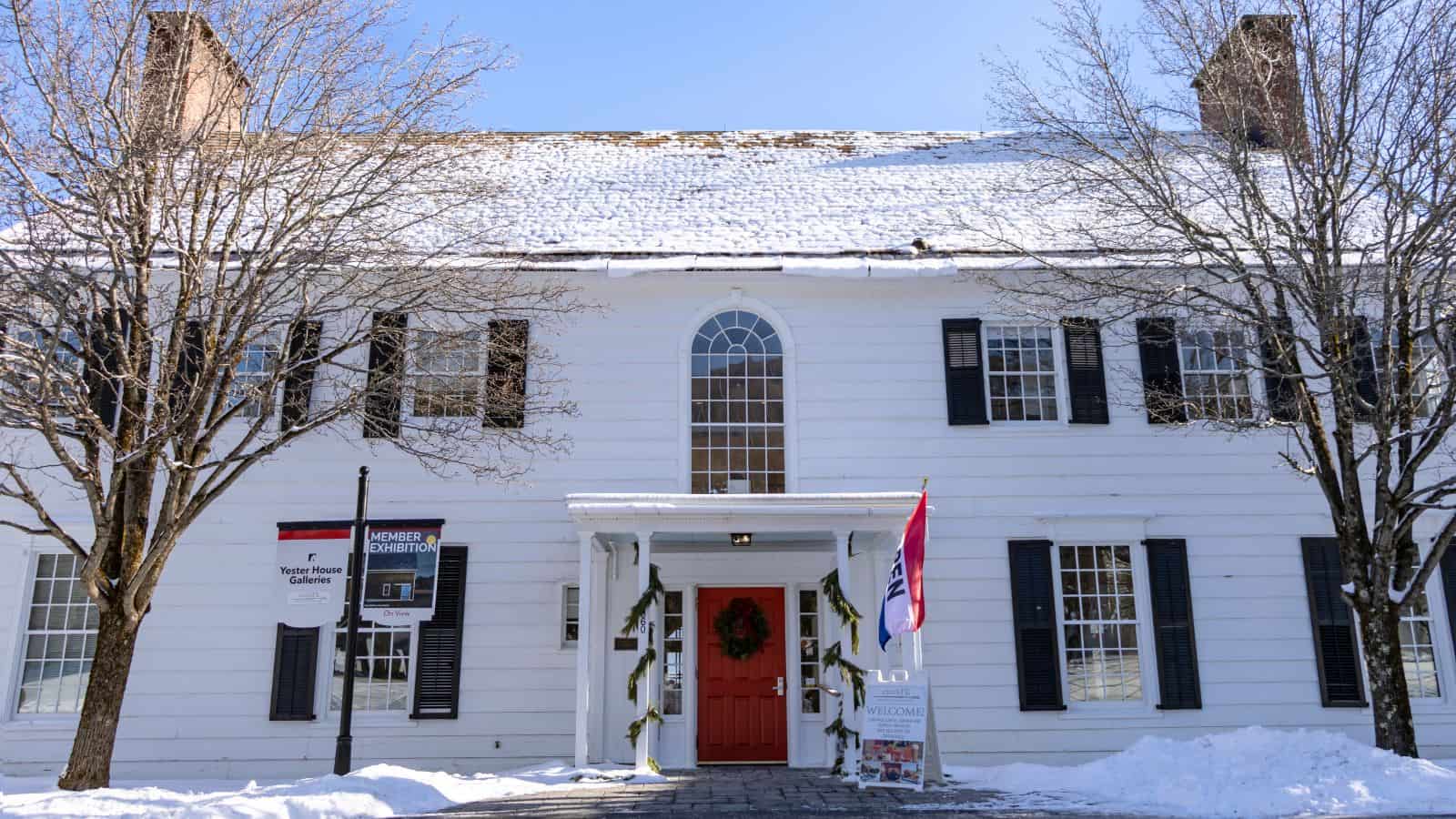 A white two-story colonial building with black shutters and a red door, snow on the roof and ground, trees at the entrance, and museum signs by the walkway.