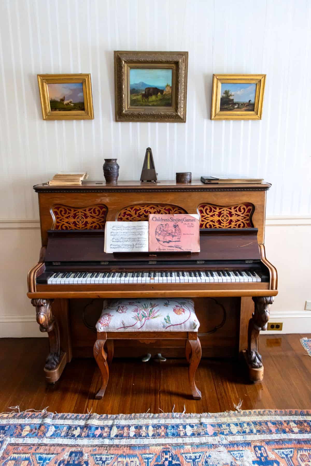 An upright piano with sheet music on its stand, a cushioned bench in front, and framed paintings and a metronome on the wall above.