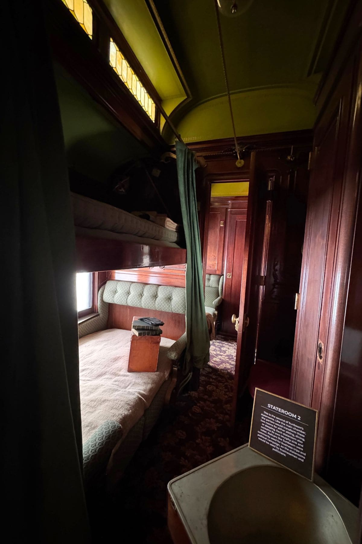 Narrow train sleeper cabin with bunk beds, wooden paneling, a small sink, a vintage suitcase, and sign labeled "Stateroom 3" in dim lighting.