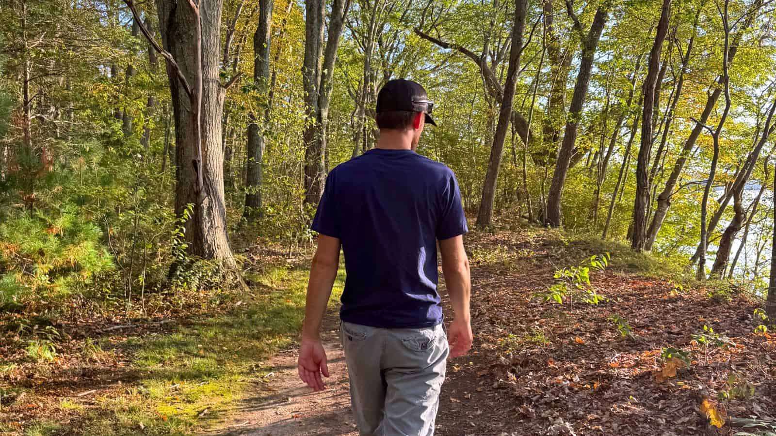 A person wearing a navy shirt, gray pants, and a cap walks along a dirt trail while hiking in New London County, Connecticut’s sunlit, leafy forest.