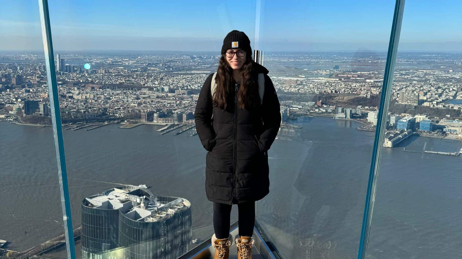Person wearing a black coat and beanie stands on a glass observation deck overlooking a cityscape and river.