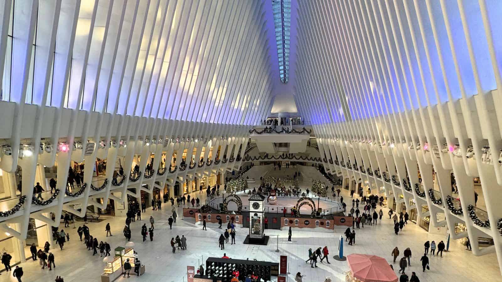 Wide-angle view of people walking inside the spacious, modern Oculus transportation hub with high white ribbed ceilings and holiday decorations.
