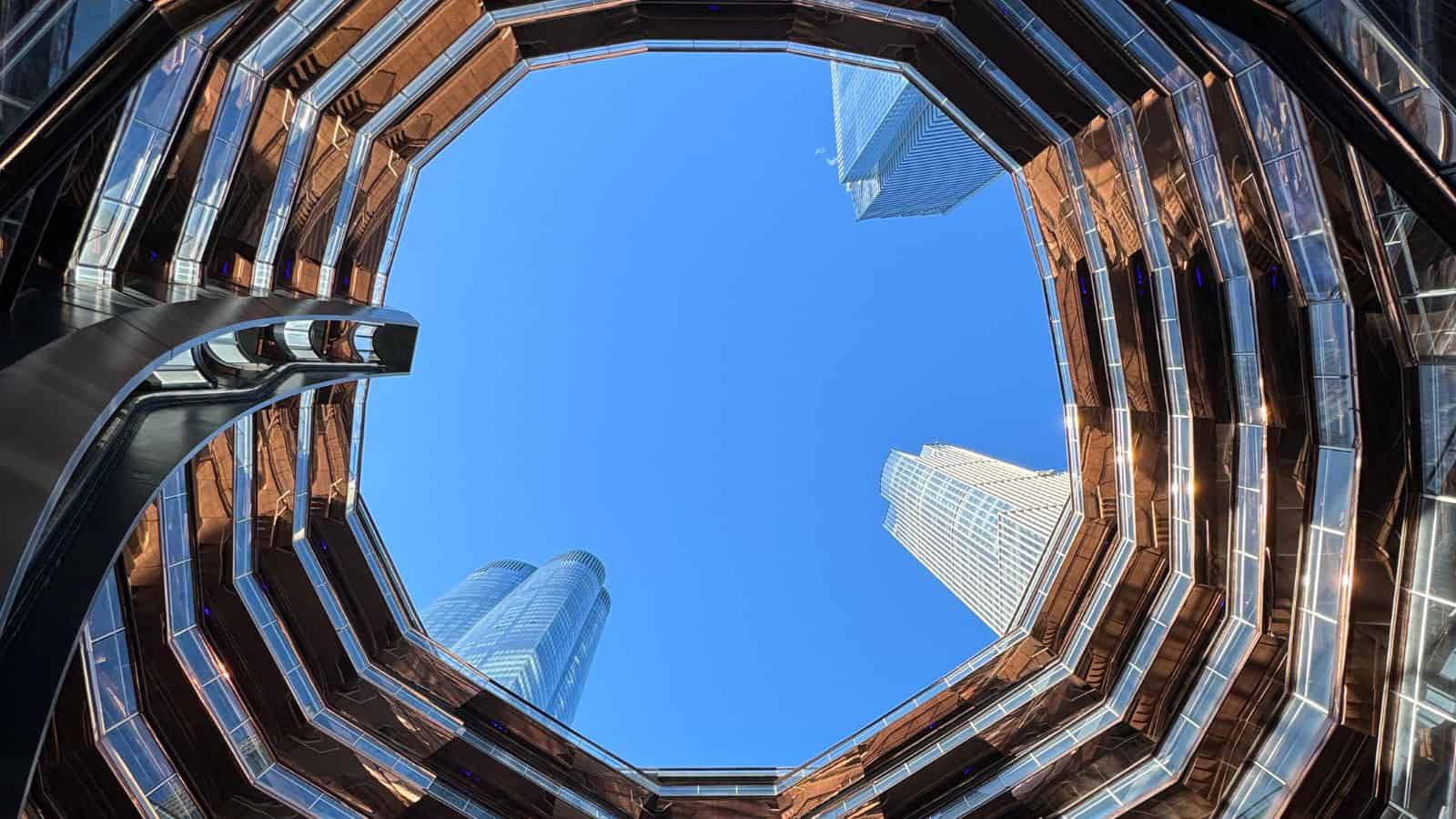 View looking up from the center of a circular, multi-level structure with modern skyscrapers visible against a clear blue sky.