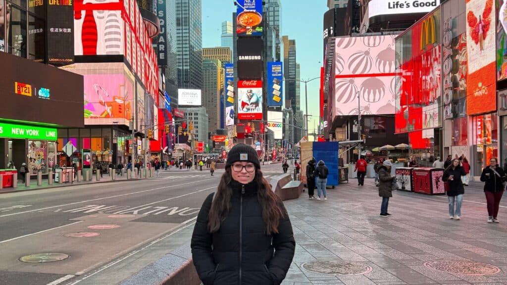 A person wearing a black winter coat and hat stands in Times Square, New York City, surrounded by bright billboards and a few pedestrians.