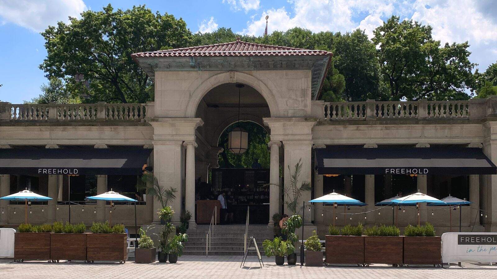 A stone building with arched entryway and two black awnings labeled "FREEHOLD." Outdoor seating with blue umbrellas and potted plants is visible in front. Trees in the background.