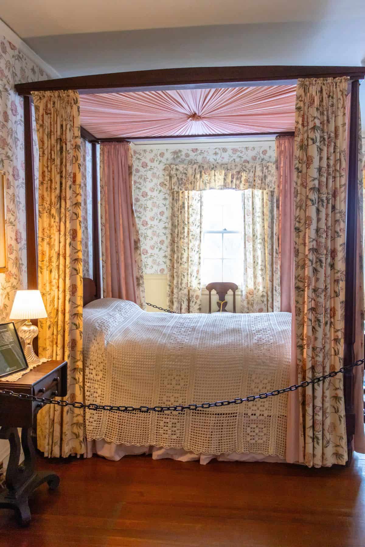 A four-poster bed with floral curtains and a crocheted bedspread stands in a vintage room with floral wallpaper and a window in the background.