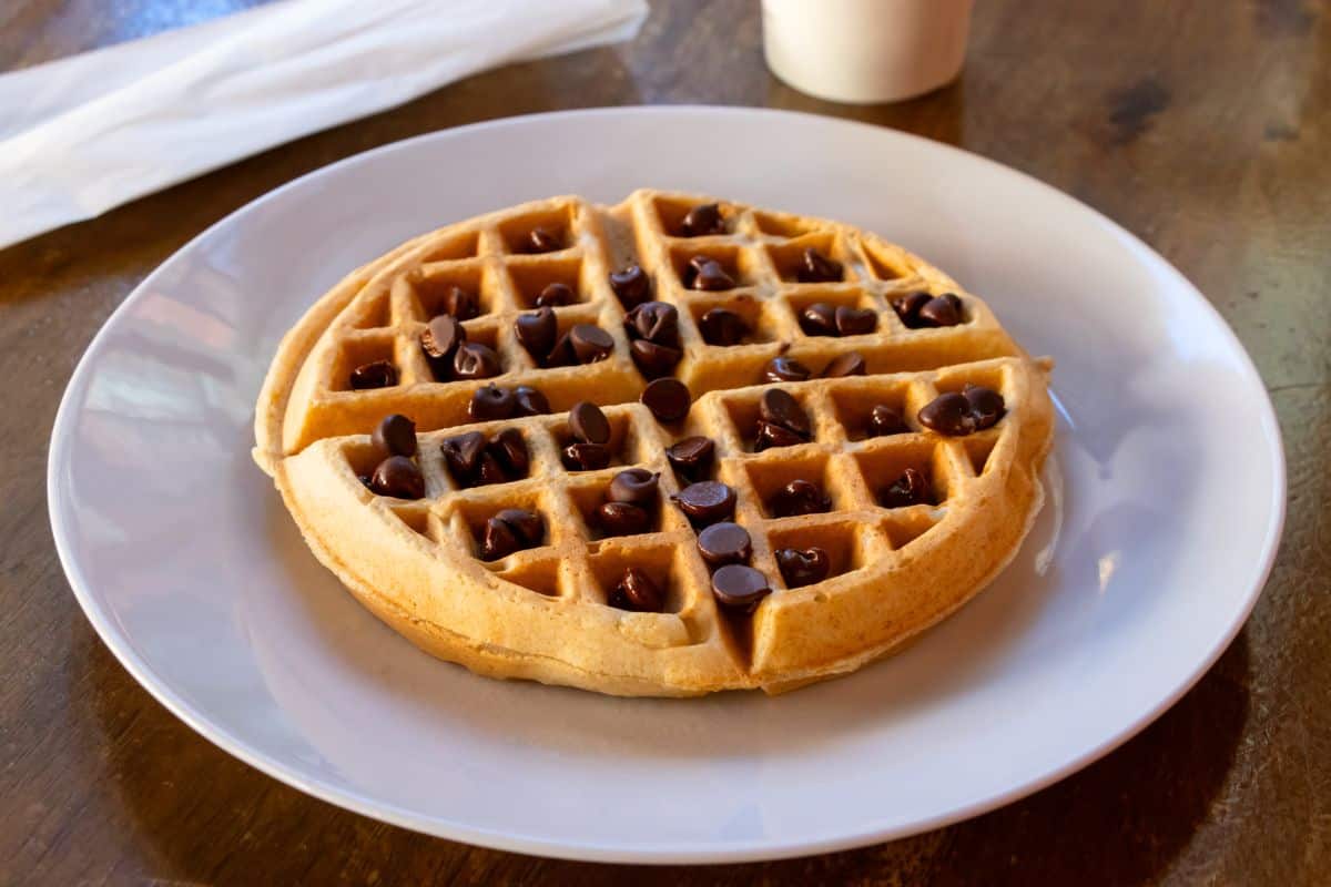 A round waffle topped with chocolate chips sits on a white plate on a wooden table.