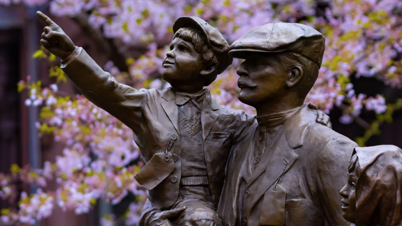 Bronze statue of a man holding a boy who is pointing, with another child beside them; cherry blossom tree in the background.