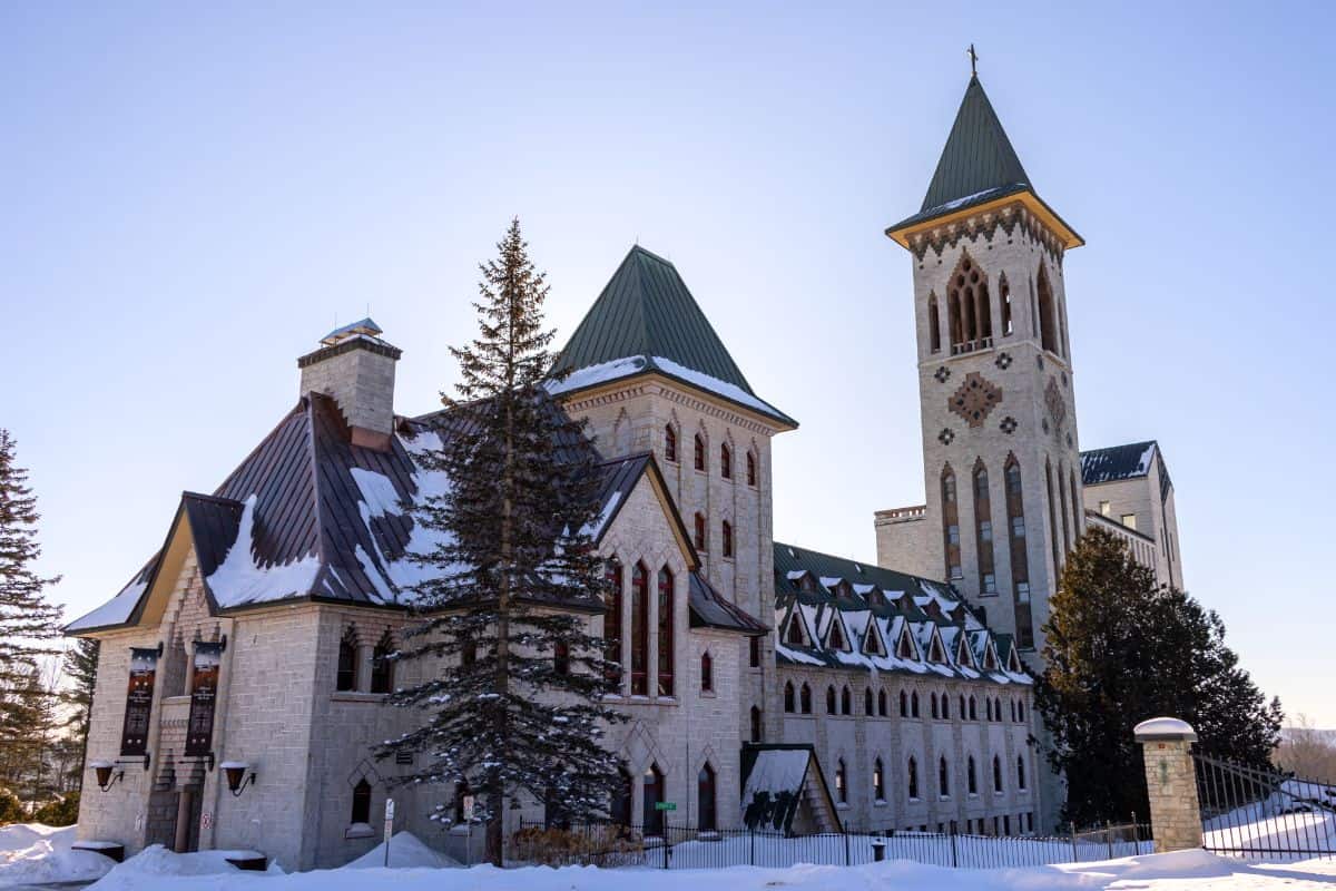 A large stone building with a tall bell tower and steep green roofs, surrounded by snow and evergreen trees under a clear sky.