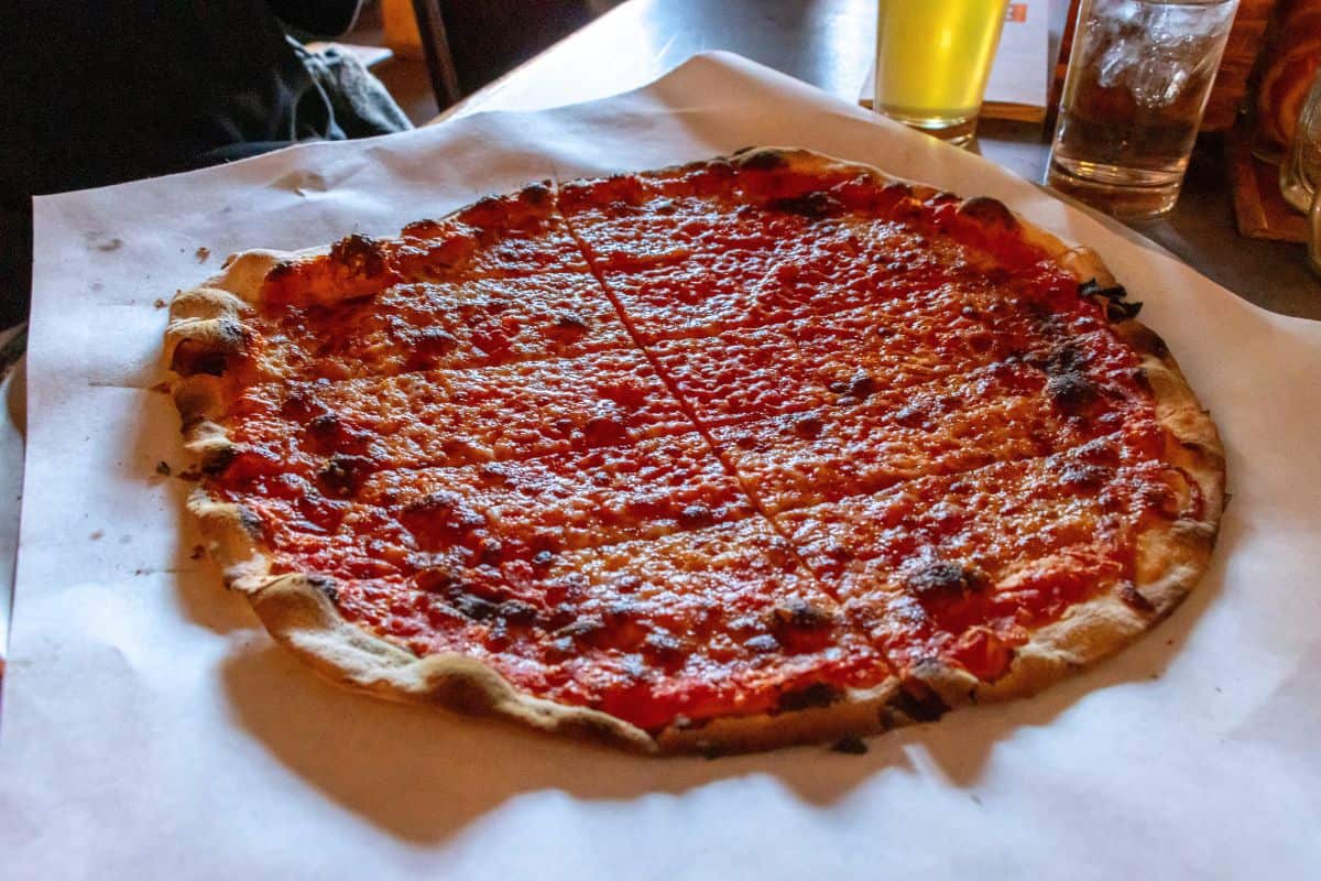 A thin-crust pizza topped with tomato sauce and minimal cheese, cut into square slices, sits on parchment paper on a table.