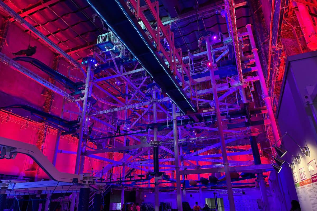 Large indoor structure with interconnected platforms, beams, and ropes, illuminated by blue and pink lights, resembling an adventure climbing course.