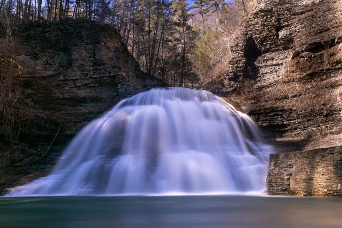 Wide waterfall cascading down rocky cliffs into a calm pool, surrounded by trees and layered stone formations under a clear sky.