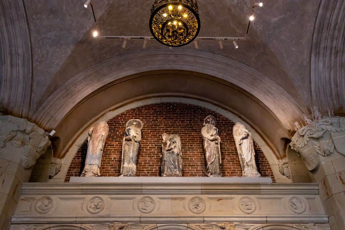 Four stone statues of robed figures stand on a ledge against a brick wall, beneath an arched ceiling with a decorative circular chandelier.