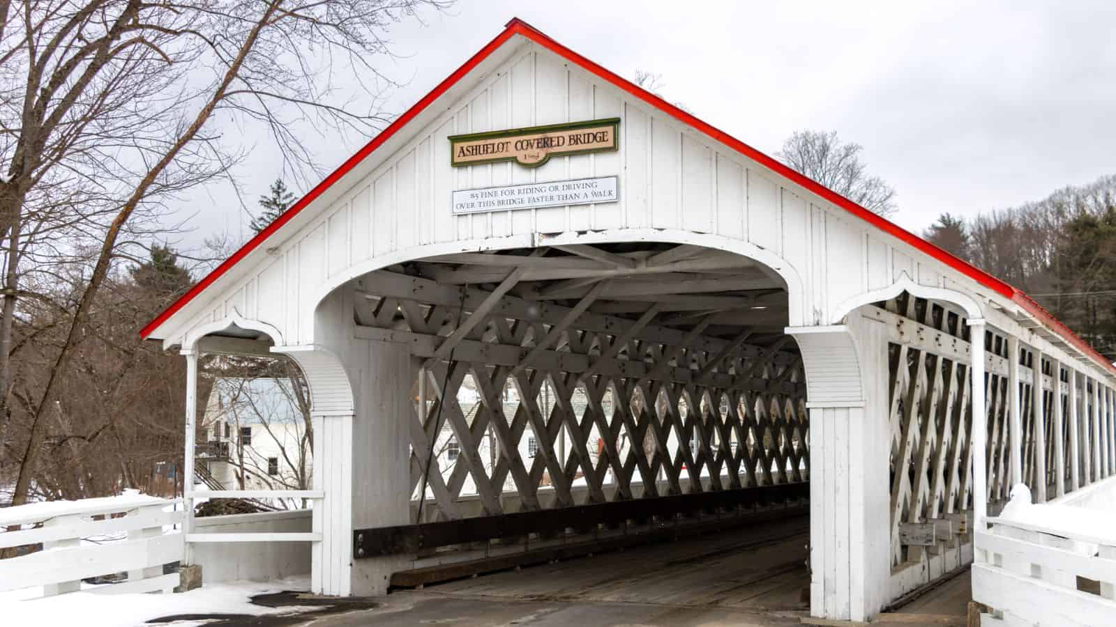 A white wooden covered bridge with a red roof, displaying a sign reading "ASHUELUT COVERED BRIDGE" and instructions above the entrance. Trees and buildings are visible in the background.