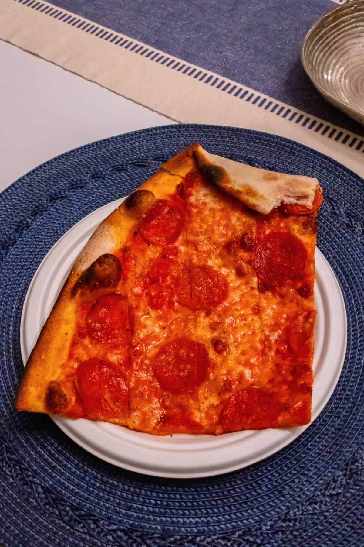 A slice of pepperoni pizza with a slightly folded crust is served on a white plate, placed on a blue woven placemat.