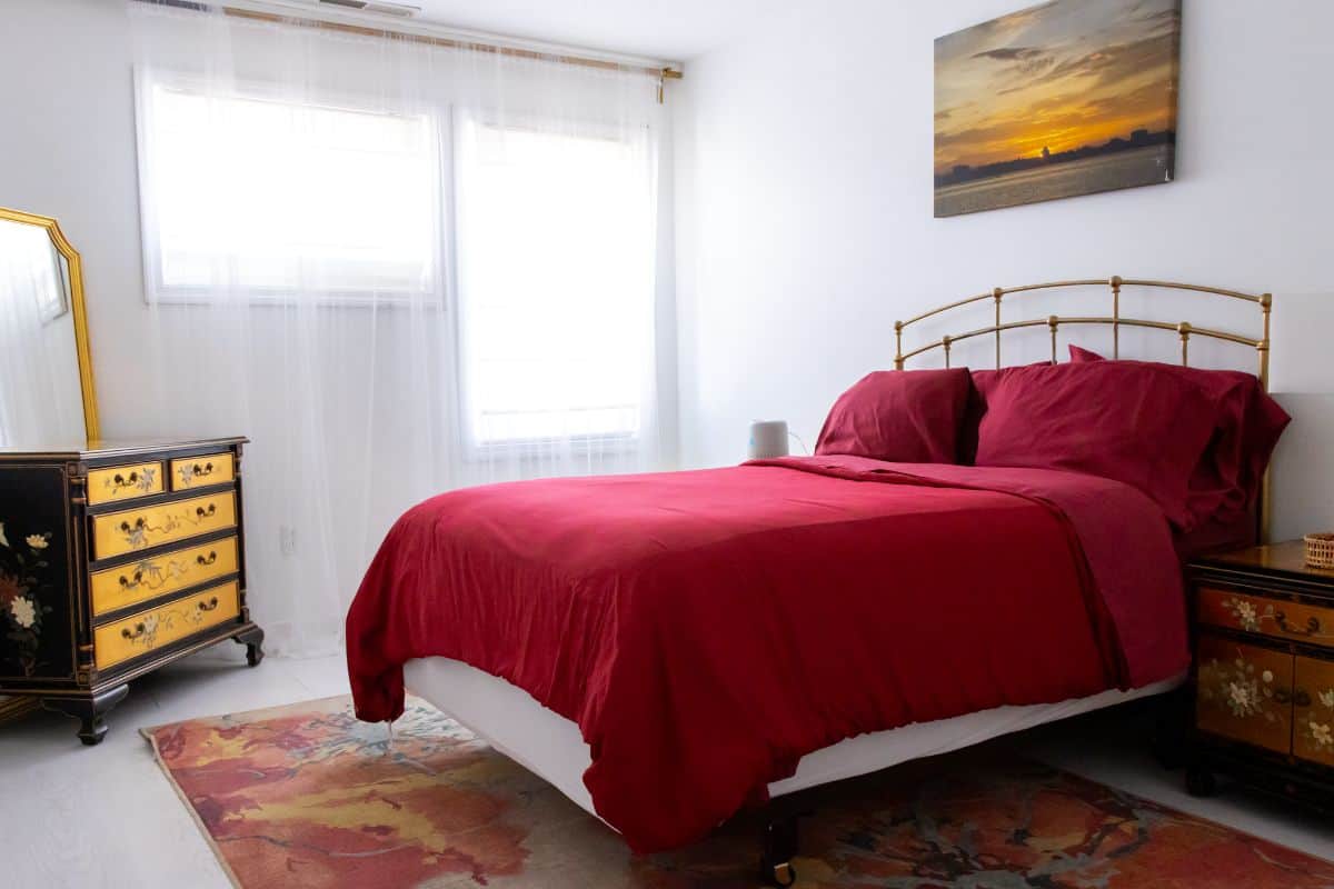 A neatly made bed with red bedding is centered in a bright bedroom. There are two windows, a floral rug, vintage dressers, and a sunset painting on the wall.