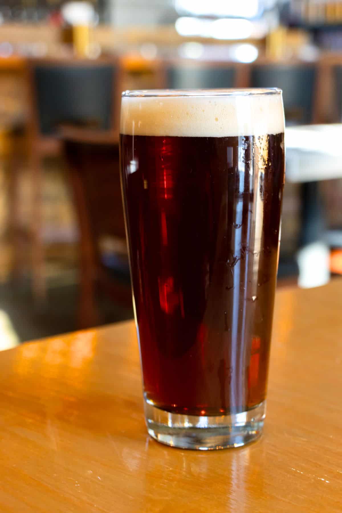 A pint glass filled with dark beer sits on a wooden table, with a blurred bar setting in the background.
