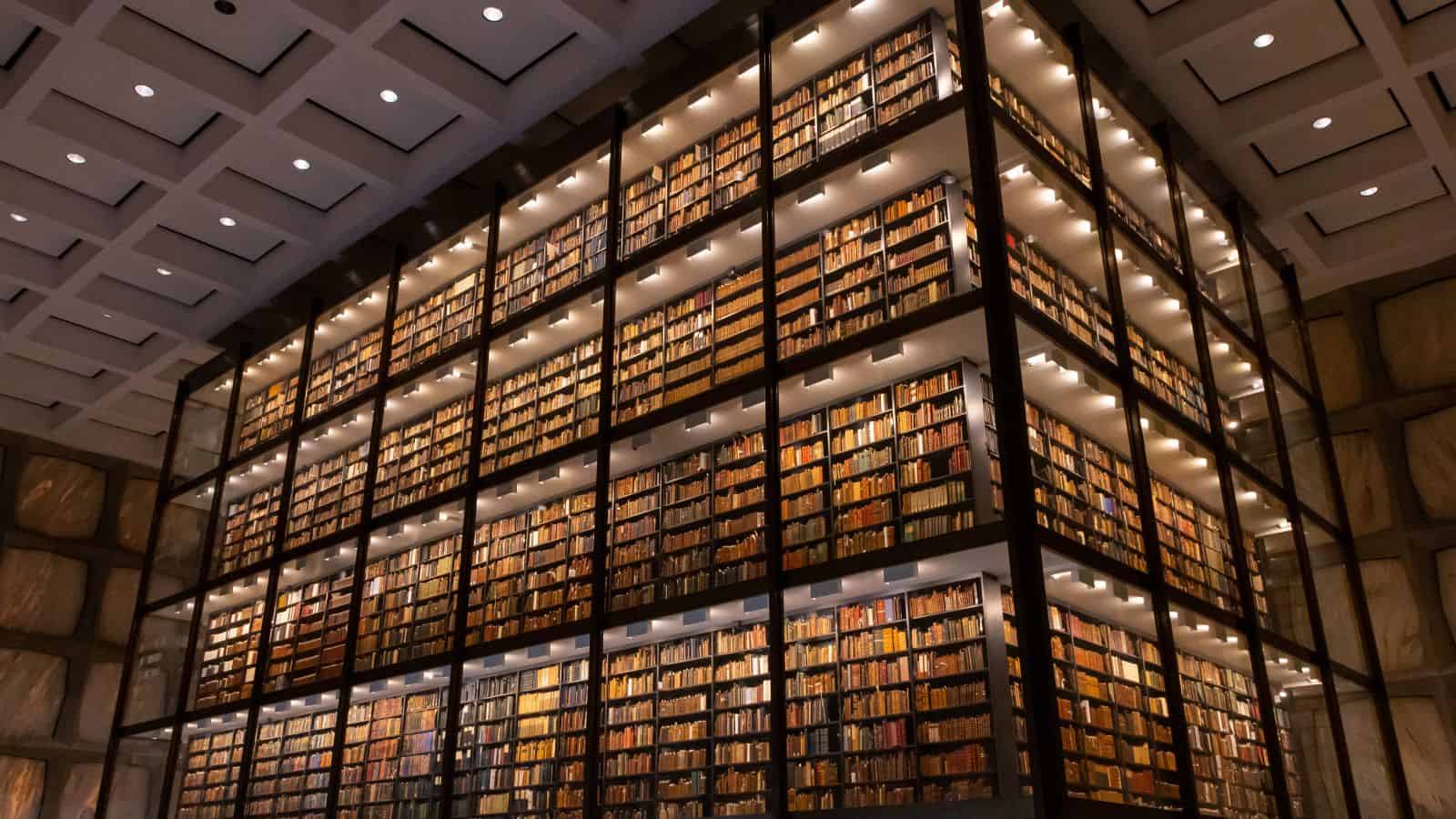 A large, multi-story glass-enclosed bookshelf filled with books stands in the center of a modern library with a patterned ceiling.