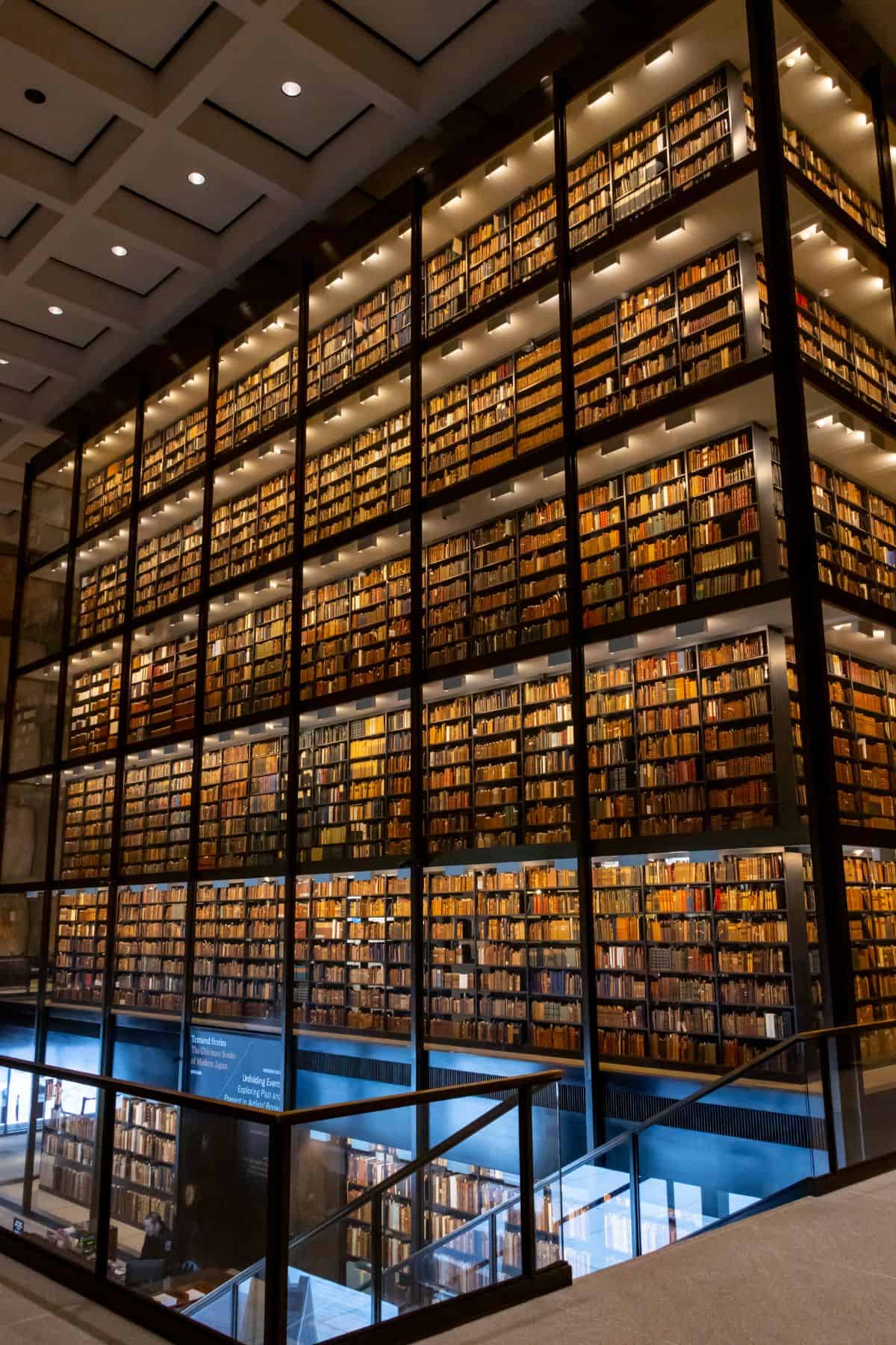 A multi-story glass-walled library stacks thousands of books across several floors, viewed from an interior balcony with modern lighting.