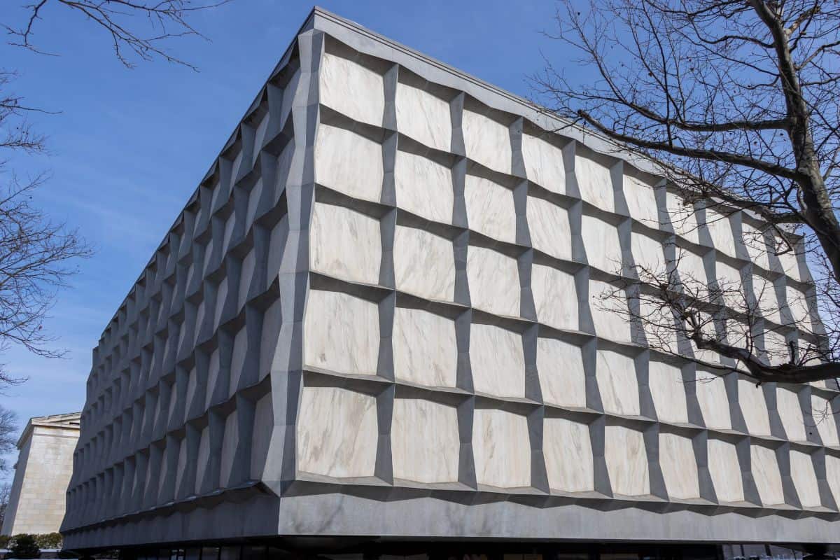 A large, windowless, cube-shaped building with a grid pattern exterior, under a blue sky, with leafless tree branches in the foreground.