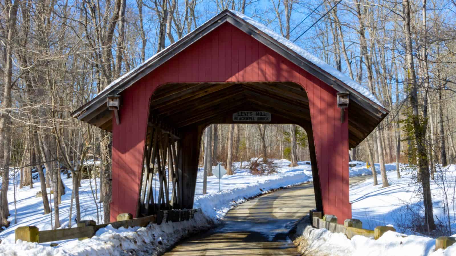 A red wooden covered bridge spans a snow-lined road through a forest of leafless trees on a sunny winter day.