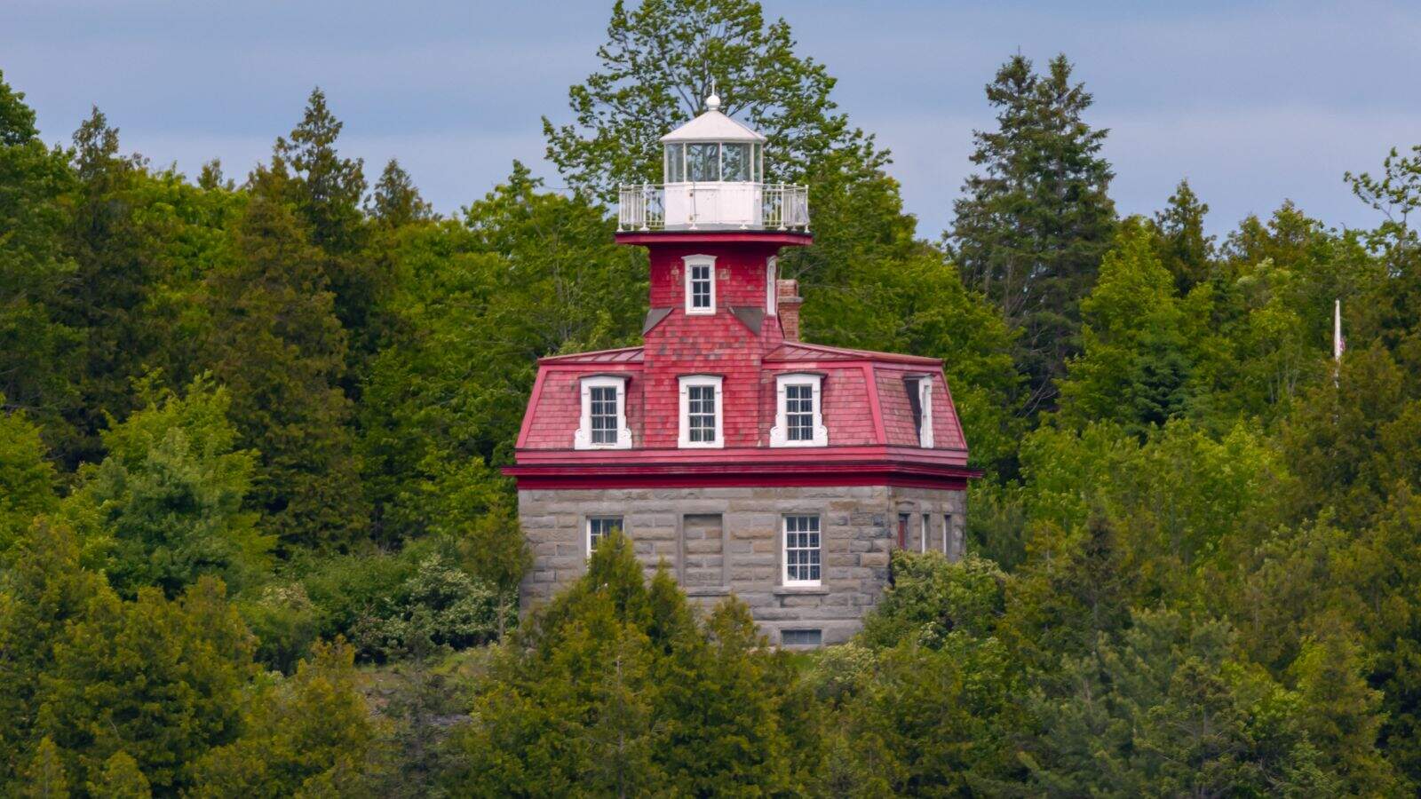 A red-roofed lighthouse with a stone base stands among dense green trees under a partly cloudy sky.