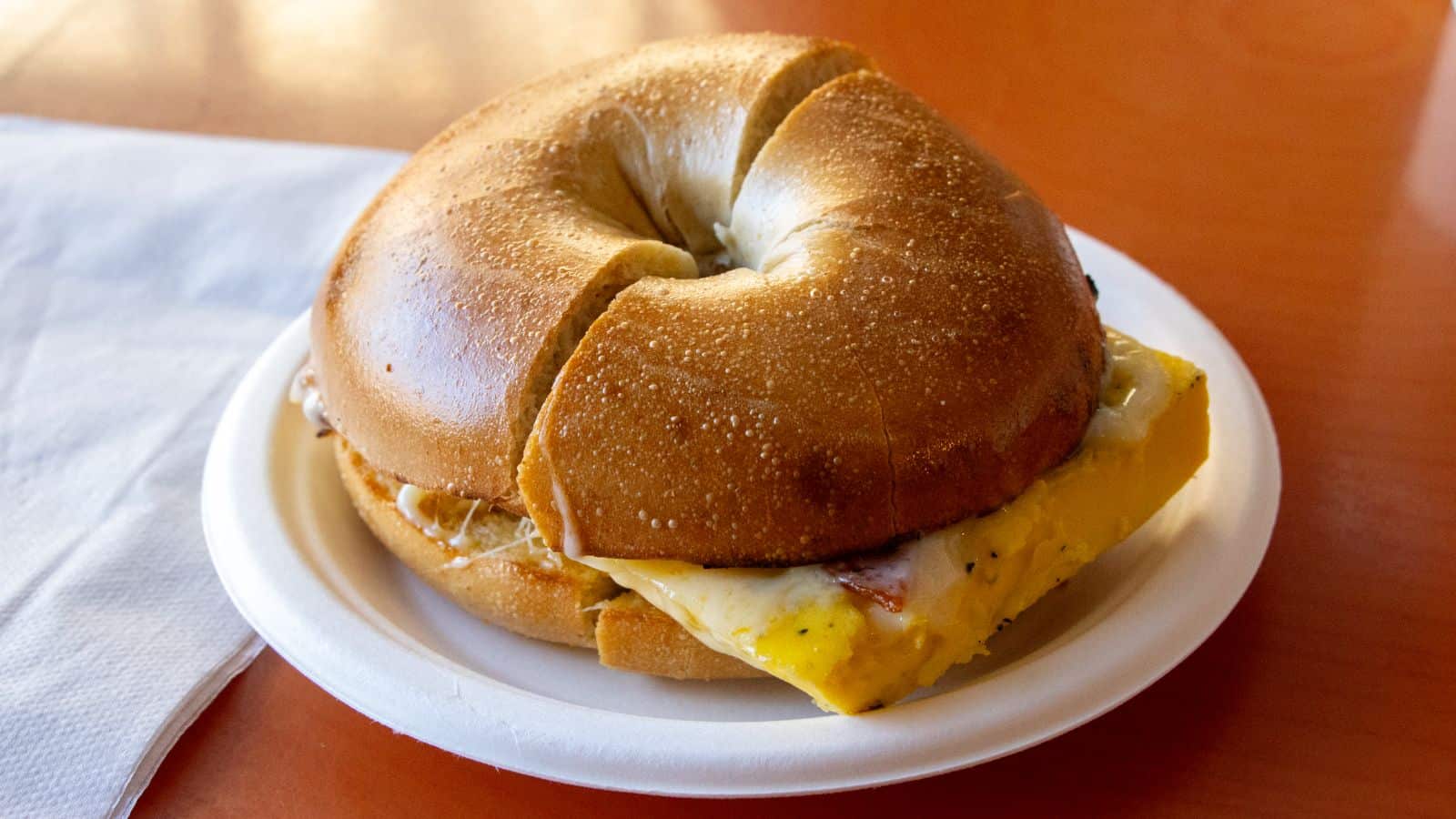 A sliced bagel sandwich with egg and cheese on a white plate, next to a folded napkin on a wooden table.
