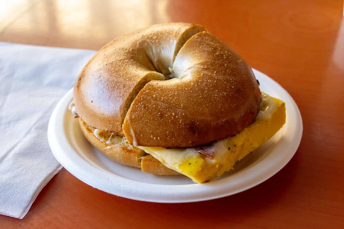 A bagel sandwich with egg and cheese on a white plate, placed on an orange table next to a folded napkin.