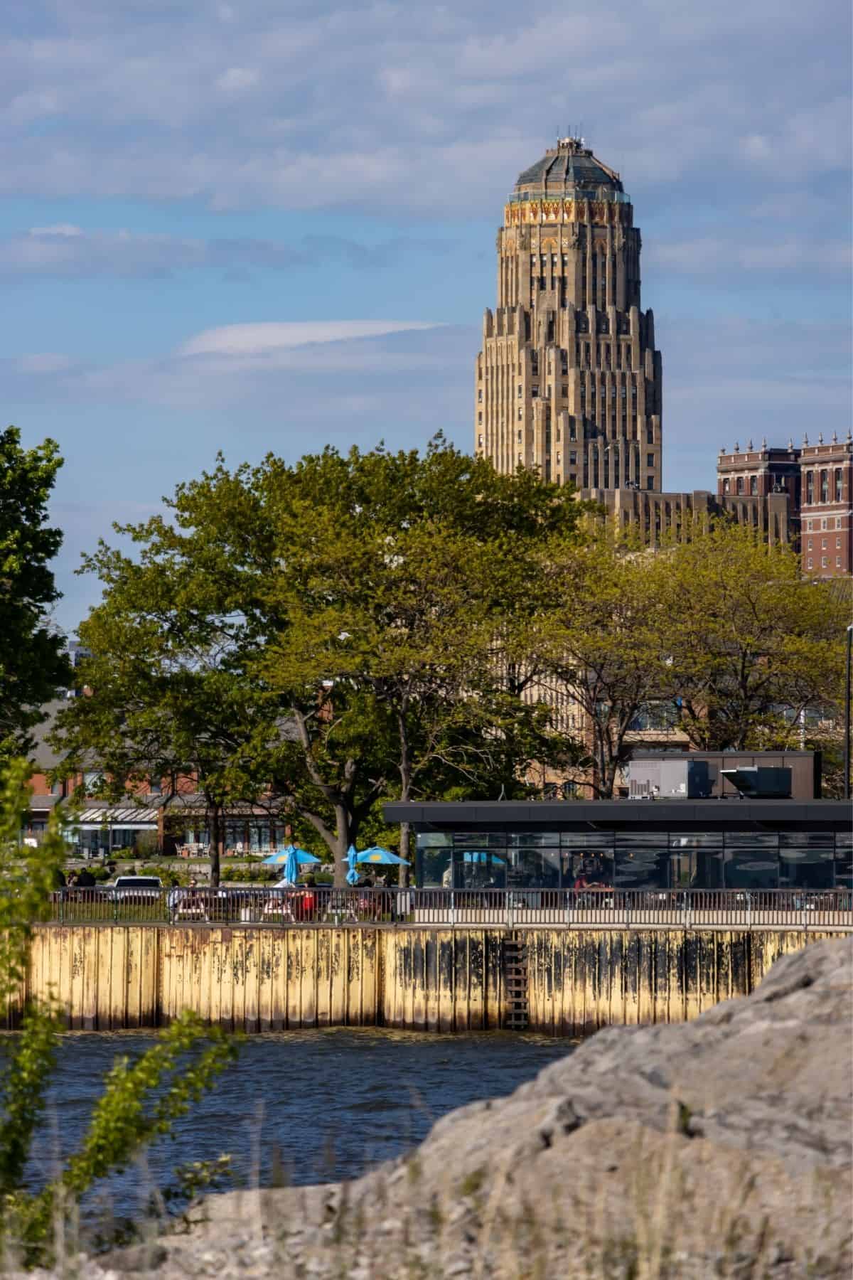 A tall art deco skyscraper rises behind trees and waterfront buildings under a partly cloudy sky.