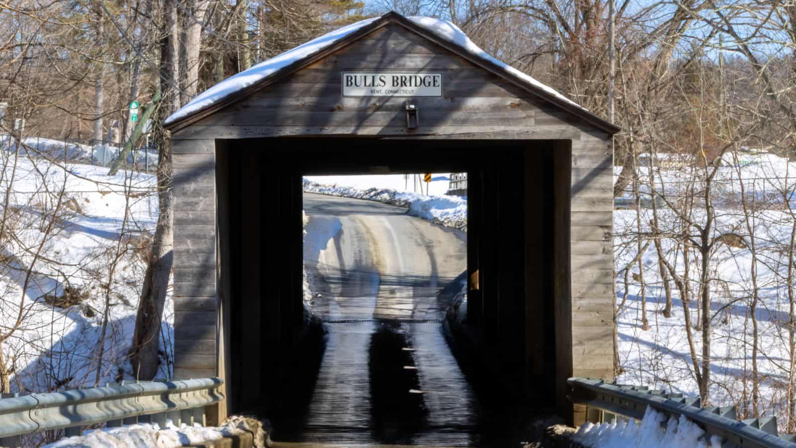 A wooden covered bridge labeled “Bulls Bridge” spans a narrow road surrounded by snow and leafless trees in winter.