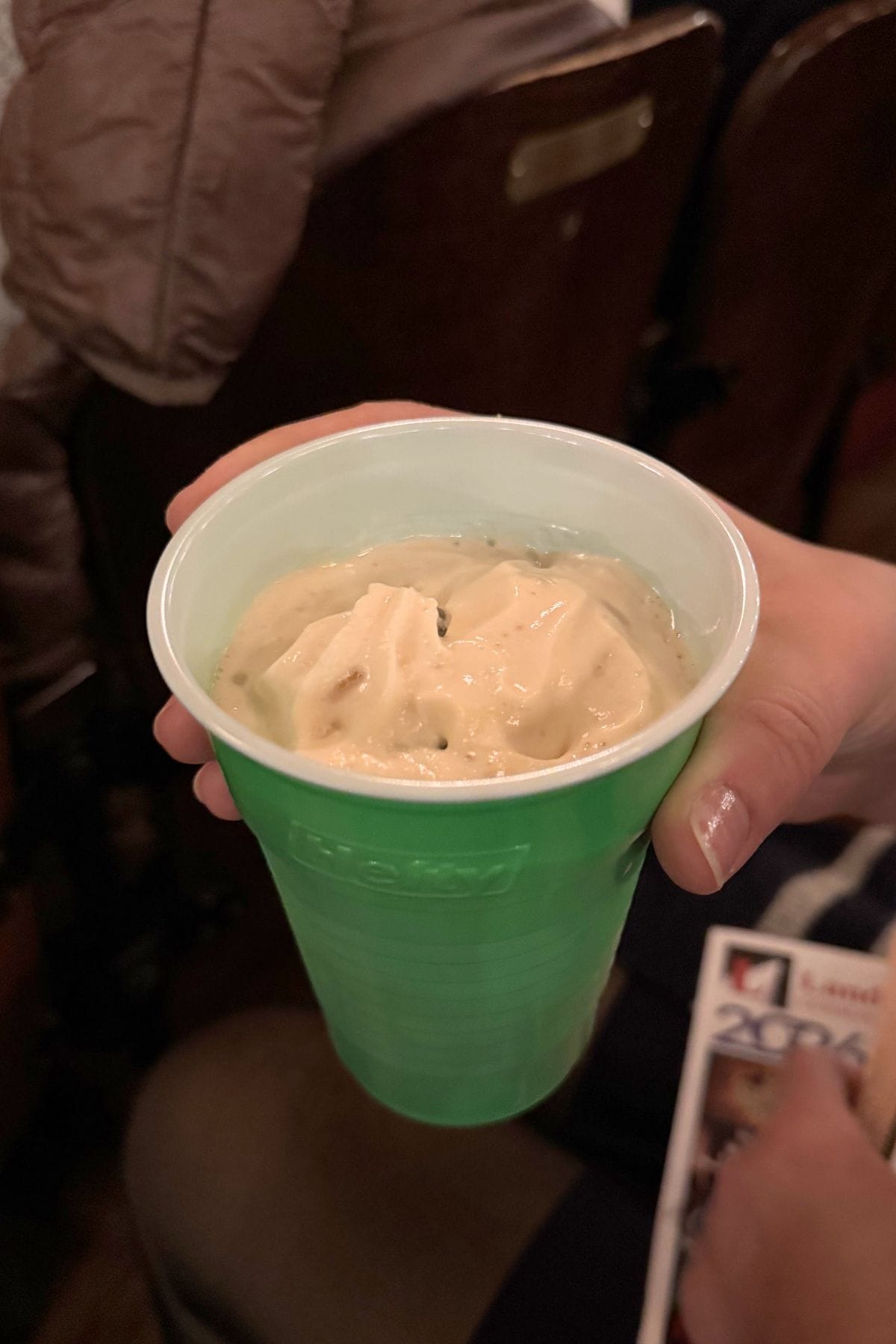 A hand holds a green plastic cup filled with a creamy, beige dessert or drink, with people seated in the background.