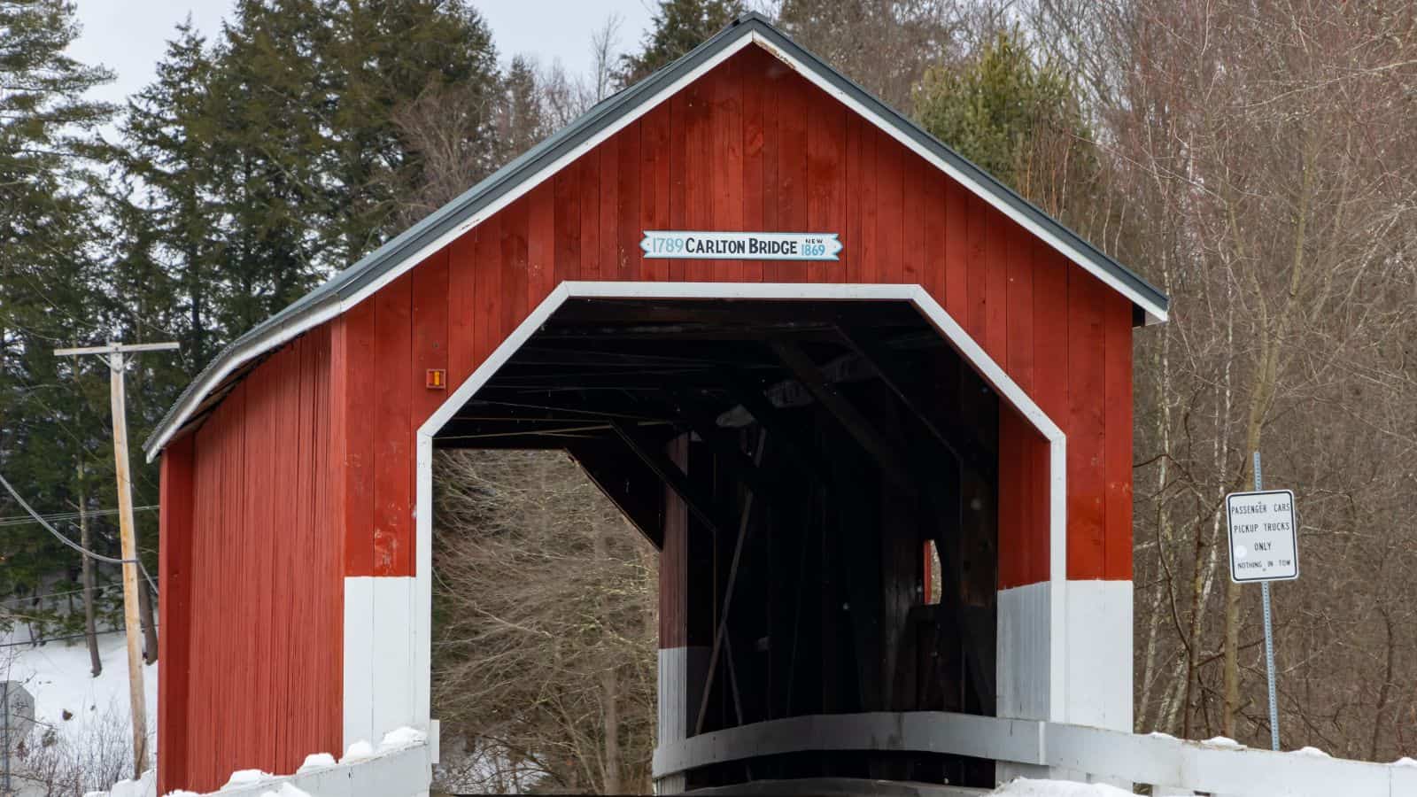 Red covered bridge labeled "Carlton Bridge" with white trim, surrounded by trees and snow, and a road sign visible on the right side.