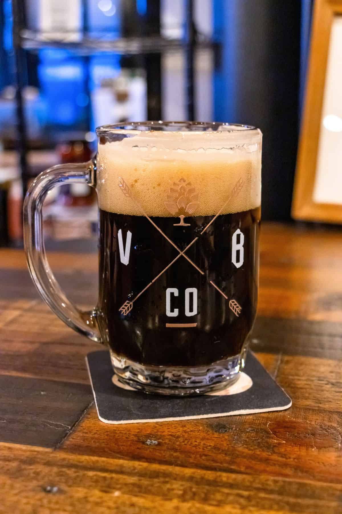 A glass mug of dark beer with a foamy head sits on a coaster atop a wooden table, displaying the logo "VB CO" on the front.