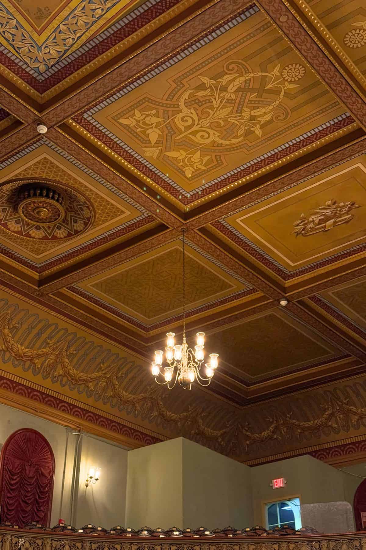 Ornate ceiling with gold and red patterns, central chandelier, and decorative walls in a theater or performance hall. Red curtains and emergency exit sign are visible.