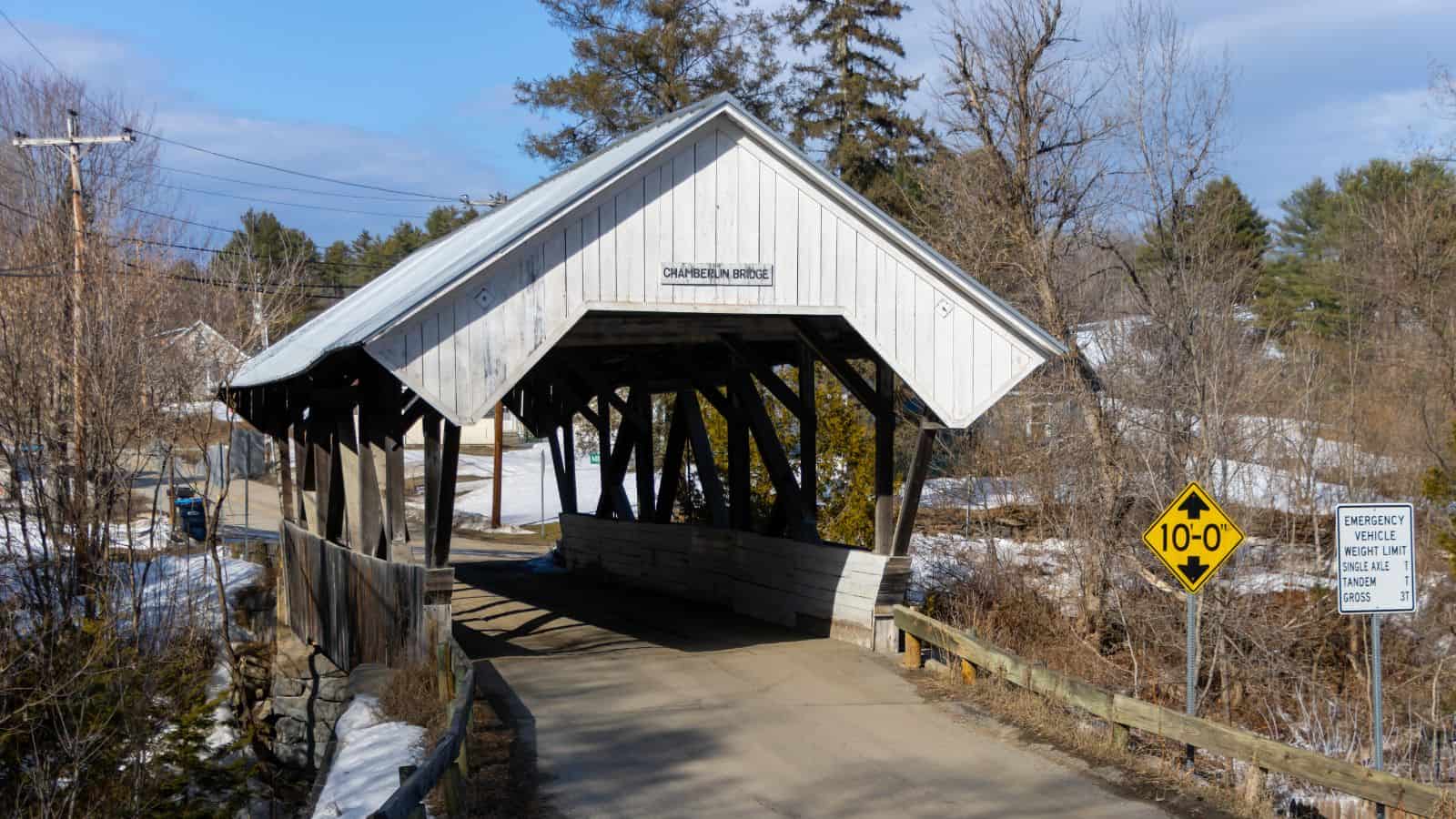 A white wooden covered bridge crosses a narrow road with a 10-foot clearance sign and snow on the ground, surrounded by trees and utility poles.