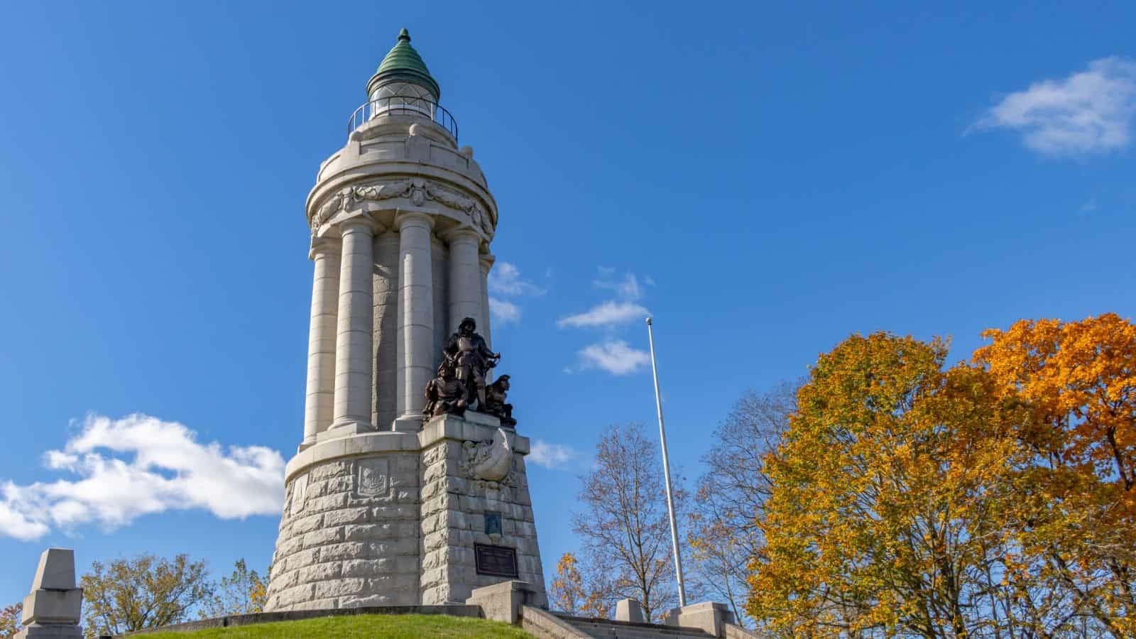 Stone monument with tall columns and a green dome, featuring a bronze statue at the base, surrounded by autumn trees under a clear blue sky.