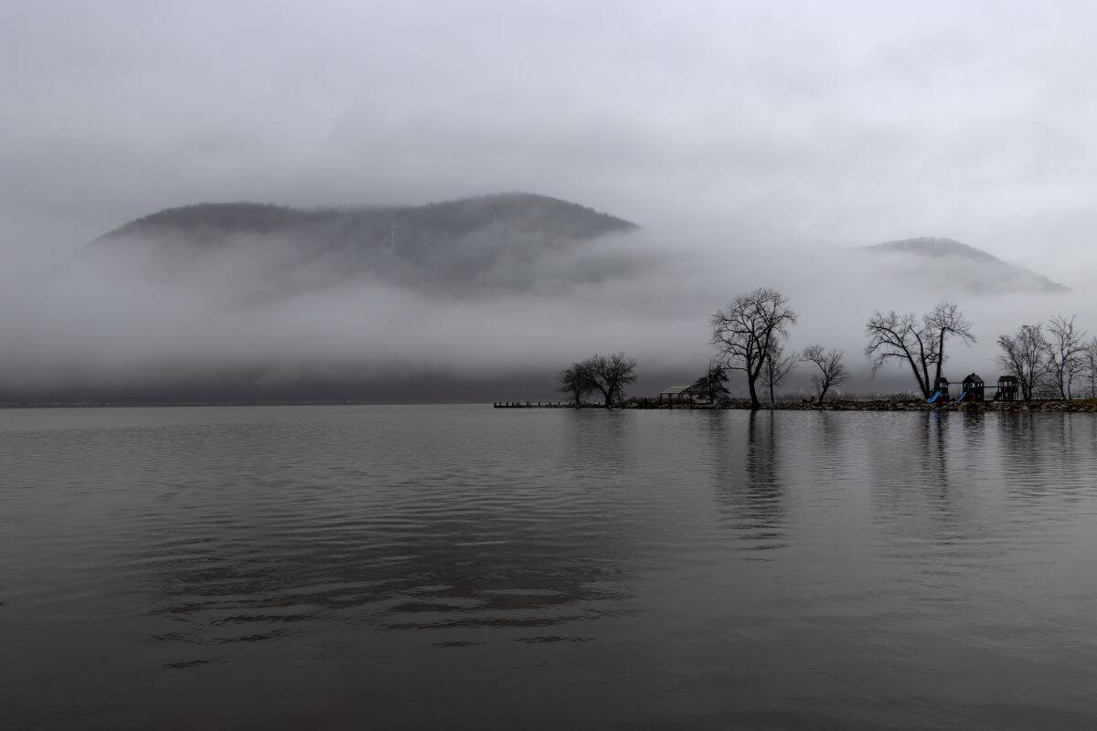 A calm lake with bare trees on the shore, low-hanging fog partially obscuring distant hills under a cloudy sky.