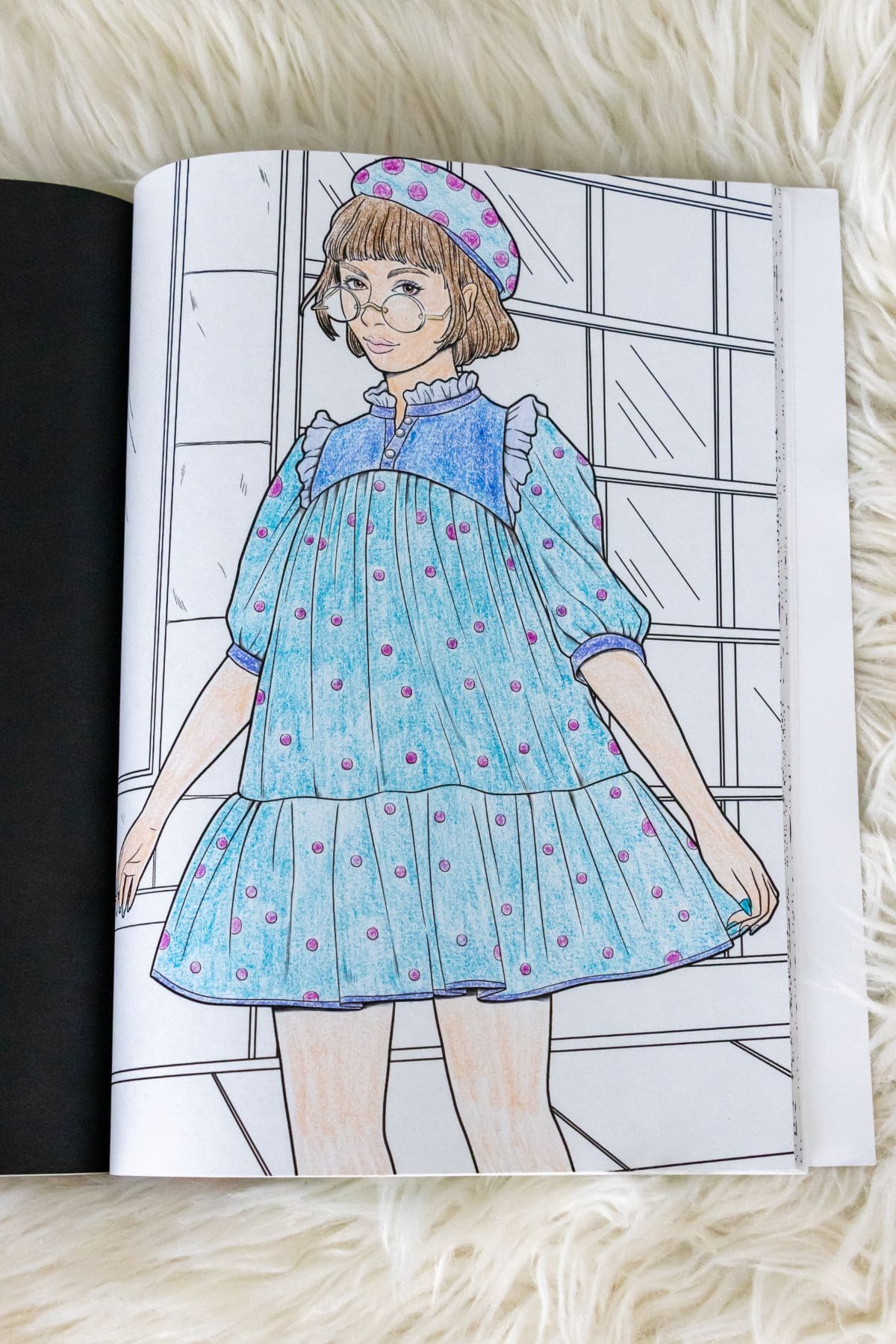 A coloring book page depicting a girl with short hair, glasses, and a beret, wearing a blue dress with pink polka dots standing in front of a window.