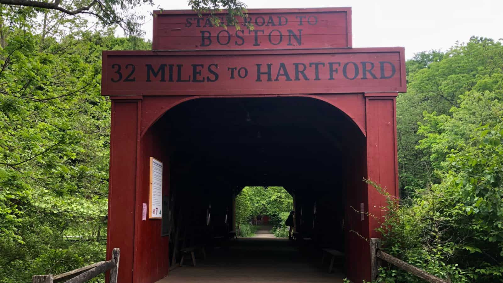 Red covered bridge with sign reading "State Road to Boston, 32 Miles to Hartford," surrounded by green trees.