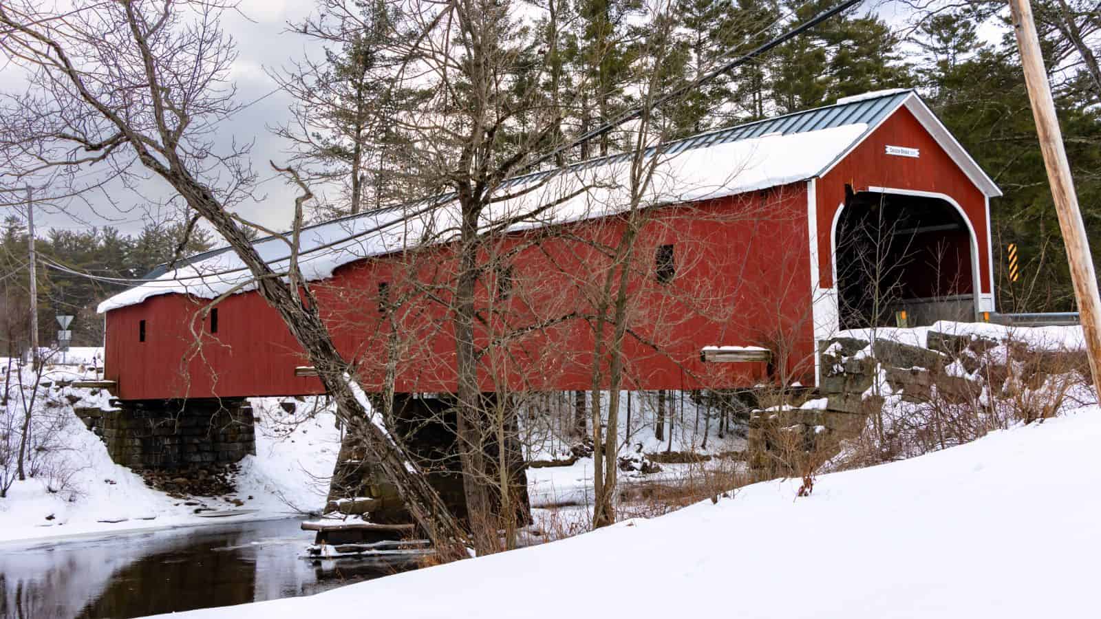 A red covered wooden bridge crosses over a small river, surrounded by snow-covered ground and bare trees in a winter landscape.