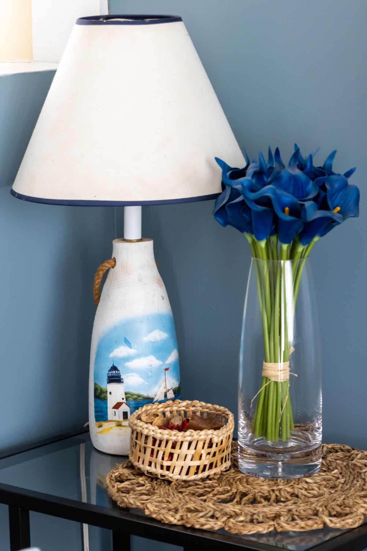 A table with a lighthouse-themed lamp, a glass vase of blue calla lilies, and a small woven basket sits against a blue wall.