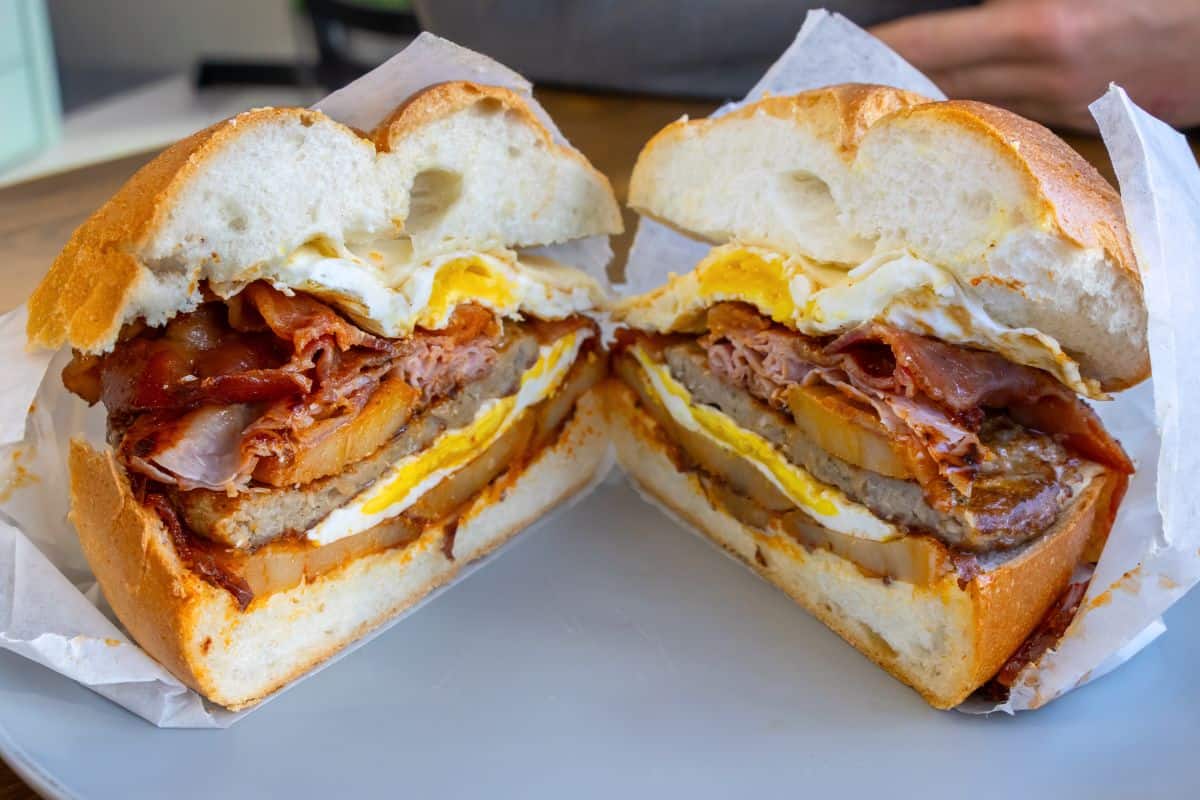 A breakfast sandwich cut in half, filled with fried egg, bacon, sausage, and cheese on a white bread roll, served on a plate.