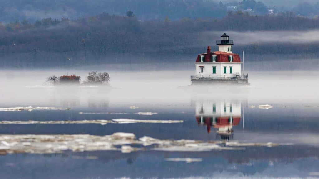 A white lighthouse with a red roof stands on a small island, surrounded by mist and calm water with a faint reflection. Trees and ice patches are visible in the background.
