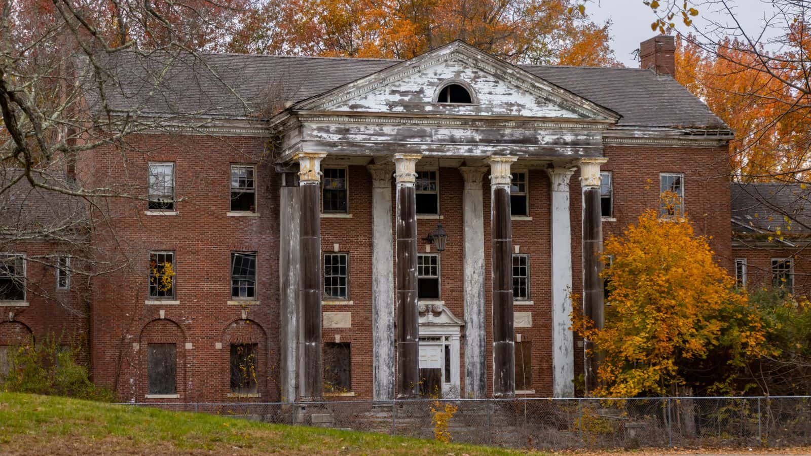 A large, abandoned brick building with tall columns and peeling paint, surrounded by autumn trees and a patchy lawn.