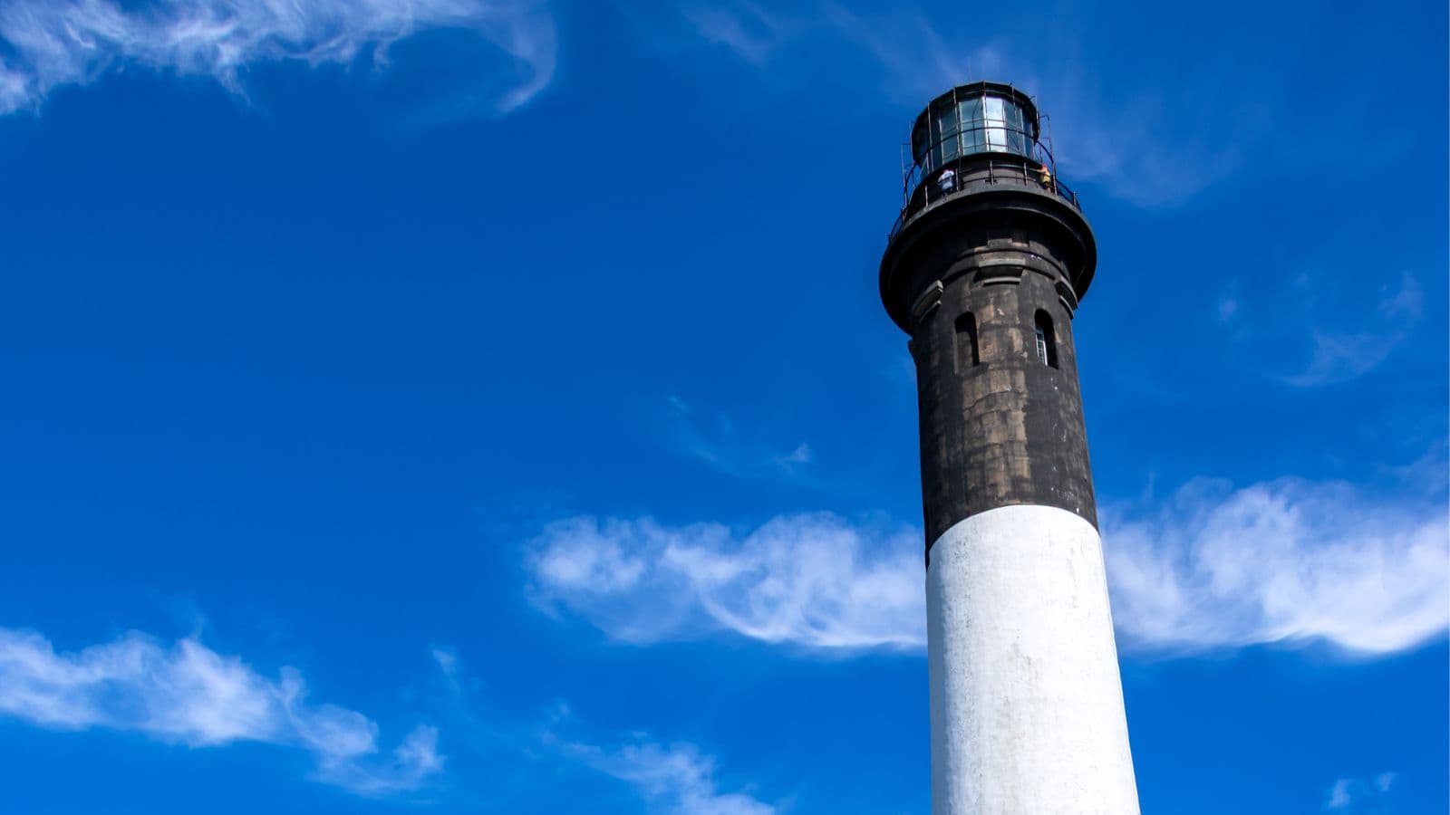 A tall lighthouse with a dark top and white base stands against a bright blue sky with thin, wispy clouds.